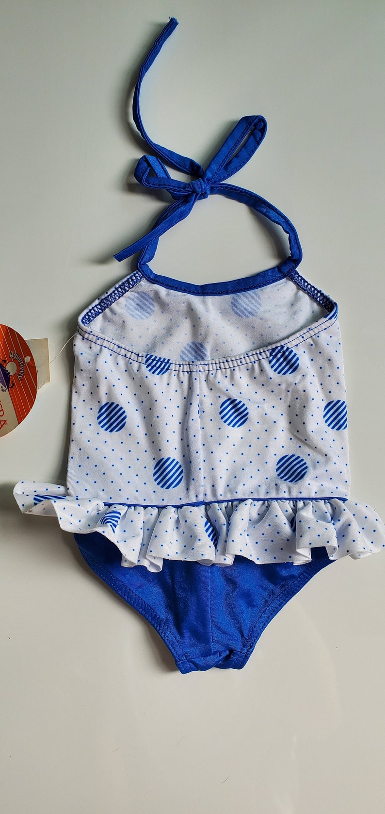 May include: A blue and white polka dot swimsuit with a ruffled bottom and a halter top. The swimsuit has a blue trim and a white background with blue polka dots.