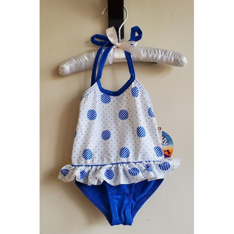 May include: A white and blue polka dot swimsuit with a blue ruffle trim and a blue halter strap. The swimsuit has a blue lining and a white tag with the text "Sun, Sand & Surf".