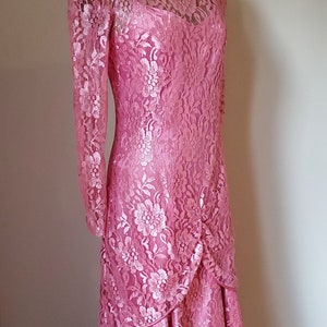 May include: A pink lace dress with a long sleeve and a high neckline. The dress has a fitted bodice and a flowing skirt with a ruffled hem.