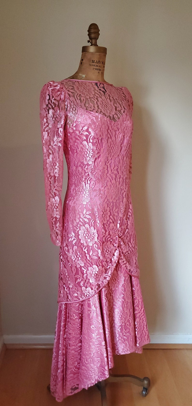 May include: A pink lace dress with a long sleeve and a high neckline. The dress has a fitted bodice and a flowing skirt with a ruffled hem.