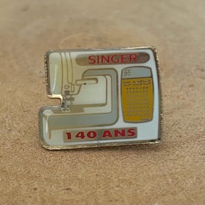 Peut inclure: Épingle à nourrice Singer représentant une machine à coudre, avec le texte "SINGER" en rouge et "140 ANS" en rouge. L'épingle est blanche, dorée et jaune.