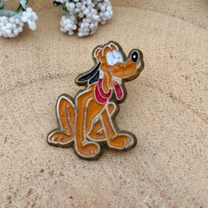 Pode incluir: Um alfinete de esmalte vintage com Pluto, o cão de desenho animado da Disney, em pose sentada. O alfinete tem uma borda dourada e é principalmente laranja com detalhes em preto, branco e vermelho. Um item de colecionador.