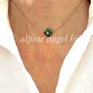 Peut inclure: Un collier délicat avec un petit pendentif en forme de trèfle à quatre feuilles vert et un motif d'éclat d'étoile doré. Le collier est porté sur une chemise blanche. Le texte "alpine angel bi" est visible.