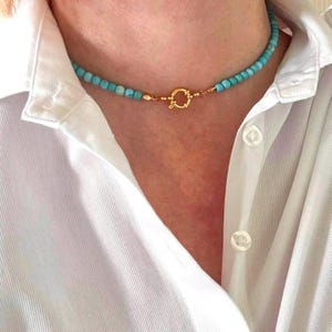Puede incluir: Un collar de cuentas turquesa con un cierre dorado. El collar está puesto en el cuello de una persona que lleva una camisa blanca.