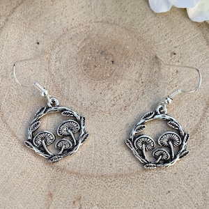 以下が含まれることがあります： 葉の輪の中に3つのキノコがデザインされた、円形のシルバー製のイヤリングです。