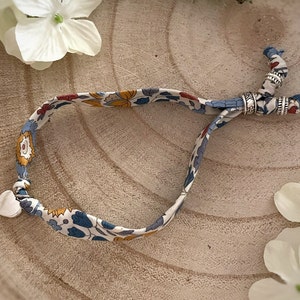 Puede incluir: Un brazalete de tela blanca con un patrón floral en azul, amarillo y rojo. El brazalete tiene un dije de corazón plateado y cuentas plateadas.