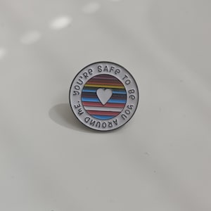 Könnte beinhalten: Runder Emaille-Ansteckpin mit dem Text "You're safe to be you around me". Der Pin hat ein weißes Herz in der Mitte, umgeben von einem regenbogenfarbenen Streifendesign.