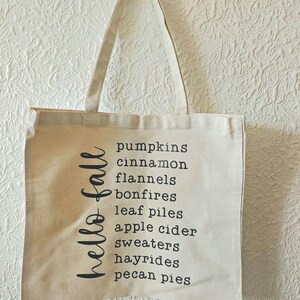 Könnte beinhalten: Eine beige Tragetasche aus Segeltuch mit zwei Henkeln, die vertikal den schwarzen Schriftzug "hello fall" sowie eine Liste mit den Wörtern pumpkins, cinnamon, flannels, bonfires, leaf piles, apple cider, sweaters, hayrides und pecan pies zeigt.