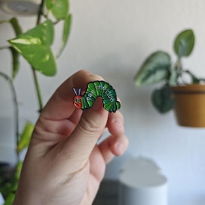 Könnte beinhalten: Ein bunter Emaille-Pin in Form einer Raupe. Die Raupe ist grün mit einem roten Kopf und den weißen Worten "eat the rich". Der Pin wird in einer Hand gehalten.
