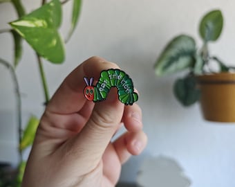May include: A green caterpillar enamel pin with a red face, purple antennae, and black outlines. The body of the caterpillar features the white text "eat the rich" written in a clear, sans-serif font across its segmented, curved back.