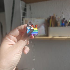 Könnte beinhalten: Ein farbenfroher Emaille-Pin in Form einer Katze. Die Katze hat ein Regenbogen-Design und eine Transgender-Pride-Flagge. Der Pin wird in einer Hand vor einem verschwommenen Hintergrund eines Regals und einer Wand gehalten.