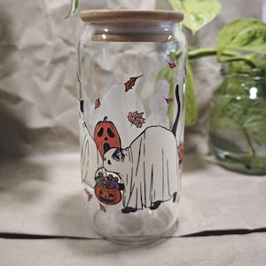 Könnte beinhalten: Ein klares Glas im Dosen-Design mit einem Holzdeckel. Der Becher ist mit einem Halloween-Motiv mit Geistern, einem Kürbis und Herbstblättern verziert. Das Design ist in Schwarz, Orange und Weiß gehalten.