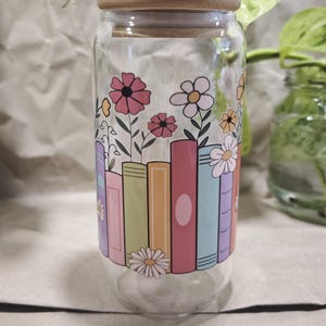 Könnte beinhalten: Ein klares Glasgefäß mit einem Holzdeckel, das mit einem farbenfrohen Design aus gestapelten Büchern verziert ist, aus denen Blumen wachsen. Die Bücher sind in den Farben Rosa, Orange, Gelb, Grün und Blau.