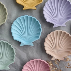Könnte beinhalten: Eine Sammlung von muschelförmigen Dekotabletts in verschiedenen Pastellfarben, darunter Gelb, Blau, Rosa und Lavendel. Die Tabletts haben eine gerippte Textur und sind auf einer grauen Oberfläche angeordnet.