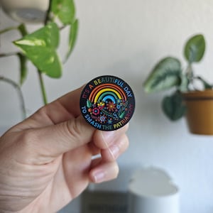 Könnte beinhalten: Ein runder, schwarzer Ansteck-Pin mit einem Regenbogen- und Blumenmuster. Der Pin trägt den Text "IT'S A BEAUTIFUL DAY TO SMASH THE PATRIARCHY" in kreisförmiger Anordnung. Das Design verwendet leuchtende Farben und detaillierte Elemente.