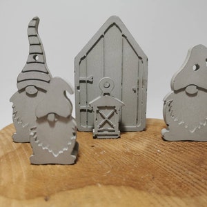 Könnte beinhalten: Drei graue, bemalte Holz-Zwerge und ein kleines, graues, bemaltes Holzhaus mit einer Tür und einer Laterne. Die Zwerge haben spitze Hüte und Bärte. Das Haus hat ein kleines Fenster und eine Tür mit einem Griff. Die Zwerge und das Haus stehen auf einer Holzoberfläche.