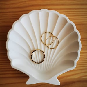 Könnte beinhalten: Weiße Keramik-Muschel-Schale mit drei goldenen Ringen darin.