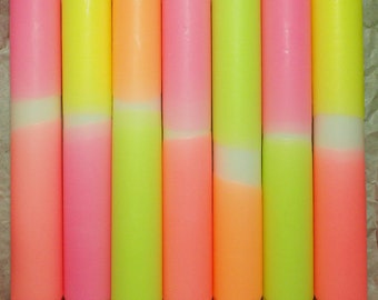 Könnte beinhalten: Sieben zylindrische Stabkerzen in einer Reihe, jede mit einem zweifarbigen Neon-Farbverlauf. Die Kerzen zeigen Kombinationen aus leuchtendem Pink, Gelb und Orange, mit einem markanten gerippten Sockel und einer spitz zulaufenden Dochtspitze.
