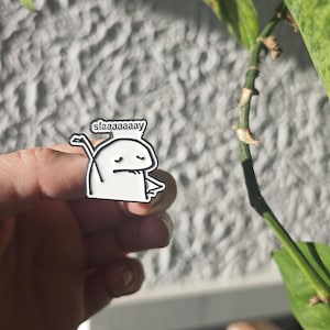 Könnte beinhalten: Ein weißer Emaille-Pin mit einer stilisierten Cartoonfigur mit dem Wort "slaaaaaaay" in einer Sprechblase. Der Pin wird in einer Hand gehalten, mit grünen Blättern und einer strukturierten Wand im Hintergrund.