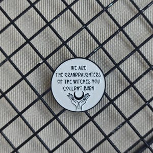 Könnte beinhalten: Runder, weißer Emaille-Pin mit schwarzem Text: „WIR SIND DIE ENKELINNEN DER HEXEN, DIE IHR NICHT VERBRENNEN KONNTET“. Der Pin zeigt einen Mond und Hände. Auf einem schwarzen Gitterhintergrund.