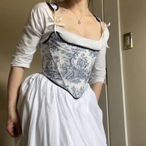 Puede incluir: Un corsé blanco y azul con estampado de toile de Jouy que representa una escena histórica. El corsé tiene un ribete azul oscuro y se lleva sobre una blusa y una falda blancas. Las correas están atadas con cintas blancas.