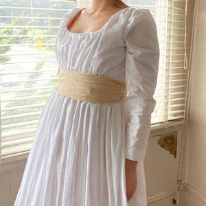 May include: A white dress with a scoop neckline and long sleeves. The dress has a gathered bodice and a fitted waist with a beige sash.