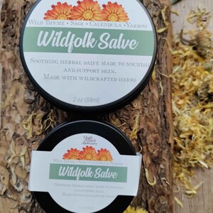May include: Two round black metal tins of Wildfolk Salve by Thistle & Peach Apothecary. The larger tin is labeled 2 oz (59ml) and the smaller tin is labeled 1 oz (30ml). Both feature white labels with orange flower illustrations and green text.