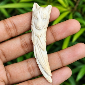 May include: An off-white, carved pendant featuring the detailed head of a wolf transitioning into a textured, feather-like design. The sculpture displays intricate lines and shading, capturing the animal's facial features and fur texture.