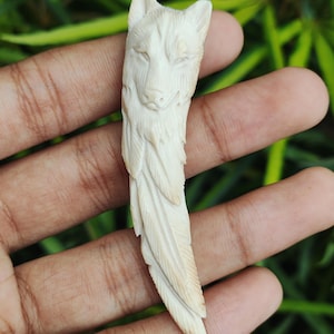 Op de afbeelding: Ivoor-kleurige gesneden hanger met een wolfskop aan de bovenkant en een veervormig ontwerp dat naar beneden loopt. Het gezicht van de wolf is gedetailleerd met vacht en gelaatstrekken. De hanger wordt in een hand gehouden tegen een vage groene achtergrond.