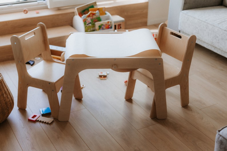 May include: A wooden table and two chairs, perfect for a child's play area. The table has a white paper roll on top, and the chairs are made of light wood.