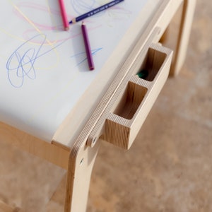 May include: A wooden children's art table with a white surface and a built-in storage drawer with three compartments. The table is made of light wood and has a simple design. There are colored pencils on the table.