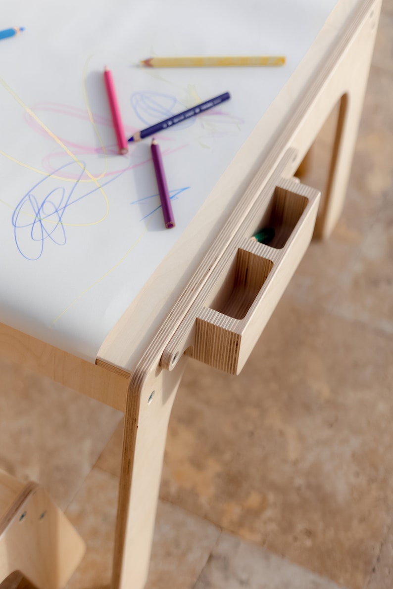 May include: A wooden children's art table with a white surface and a built-in storage drawer with three compartments. The table is made of light wood and has a simple design. There are colored pencils on the table.