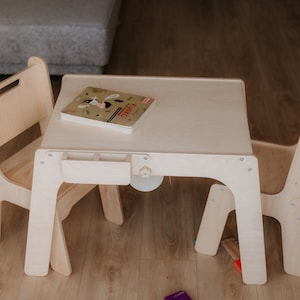 May include: A white wooden table and two chairs for children. The table has a small drawer and a book on top. The chairs are simple and sturdy.