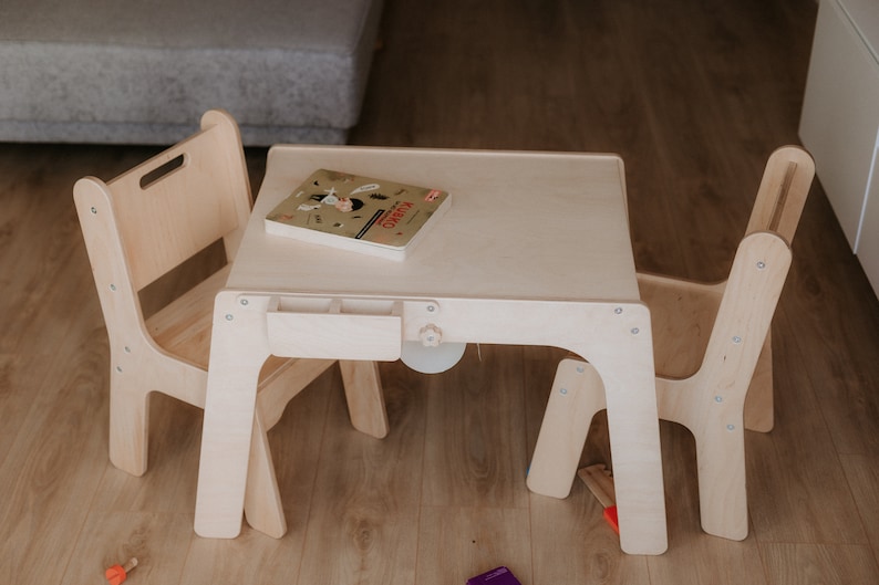 May include: A white wooden table and two chairs for children. The table has a small drawer and a book on top. The chairs are simple and sturdy.