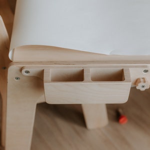 May include: A wooden children's art table with a white paper roll and a built-in storage compartment with three slots. The table has a wooden knob for adjusting the height.