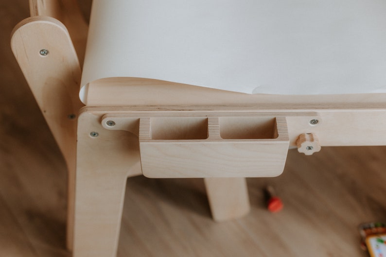May include: A wooden children's art table with a white paper roll and a built-in storage compartment with three slots. The table has a wooden knob for adjusting the height.