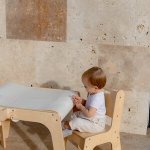 May include: A child sits at a wooden table and chair set, drawing on a roll of paper. The table has a white surface and the chair has a light wood finish.