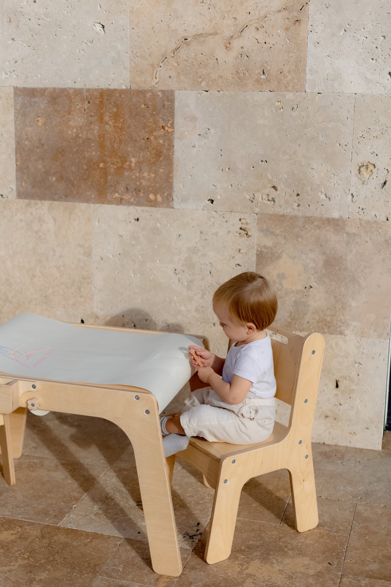 May include: A child sits at a wooden table and chair set, drawing on a roll of paper. The table has a white surface and the chair has a light wood finish.