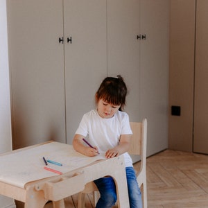 May include: A young child sits at a wooden table and chair set, drawing on a sheet of paper with colored pencils. The table has a built-in storage compartment for supplies.