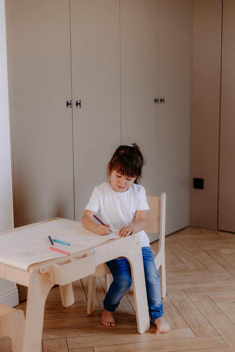May include: A young child sits at a wooden table and chair set, drawing on a sheet of paper with colored pencils. The table has a built-in storage compartment for supplies.