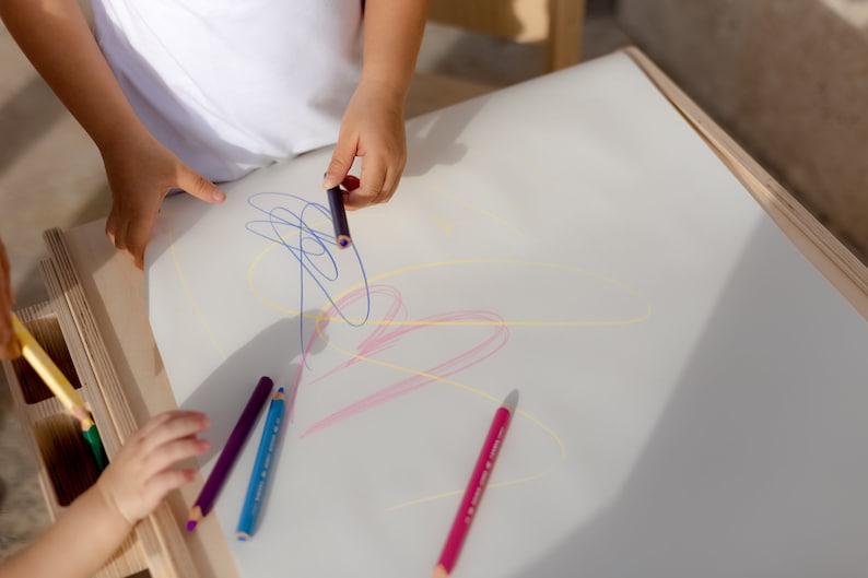 May include: Two children are drawing on a white surface with colorful pencils. The pencils are arranged on the table, and the children are using them to create lines and shapes.