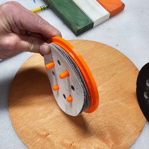 May include: An orange and gray sanding disc assembly with multiple sanding pads and orange pegs. The assembly is held over a round wooden surface. A wooden plank painted green, white, and orange is in the background.