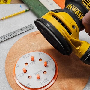 May include: A yellow and black DeWalt orbital sander is held over a wooden disc with an orange rim and a sanding disc. The sanding disc has the text "P120" printed on it. A pencil and ruler are also visible.