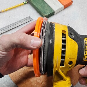 May include: A yellow and black DeWalt orbital sander with an orange sanding pad is held over a wooden surface. The image also shows a wooden piece painted green, white, and orange, along with a pencil and ruler.