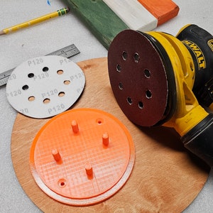 May include: A yellow and black DEWALT orbital sander with a red sanding pad, a round wooden base, and an orange 3D-printed part. Also visible are sanding discs labeled "P120" and a wooden sign with green, white, and orange sections.