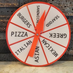 May include: A white and red spinning wheel with a red arrow pointer. The wheel is divided into eight sections, each with a different food category written in black: Burgers, Pizza, Italian, Asian, Mexican, Greek, Seafood, and Sandwiches.