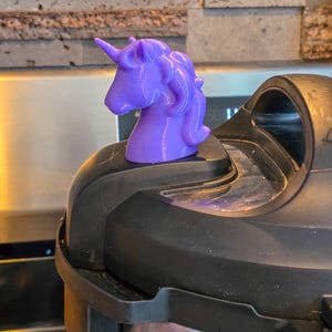 Puede incluir: Una figura de unicornio morado, con una melena y cuerno detallados, se encuentra sobre un electrodoméstico negro. La figura es de un tono morado vibrante, y el electrodoméstico tiene una base de acero inoxidable.