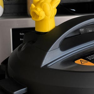 Può includere: Un deflettore di vapore per pentola a pressione a forma di drago giallo brillante. Il deflettore è montato su una pentola a pressione nera. Lo sfondo include un barattolo e un muro di piastrelle grigio scuro.