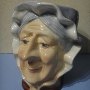 May include: A ceramic figurine of a woman's head with a white cap and a brown collar. The woman has a large nose and a kind smile.