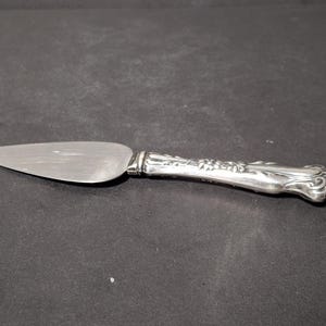 May include: A silver-toned cake server featuring a flat, triangular blade with a brushed finish and a decorative, ornate handle with embossed floral and scrollwork details. The metal handle transitions into a smooth, polished bolster near the blade.